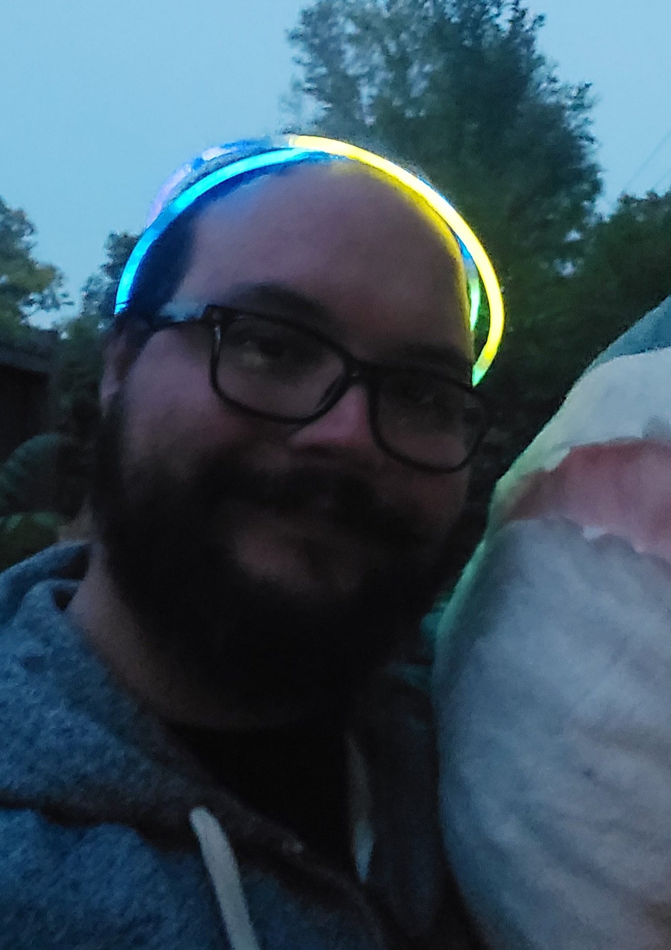 A man with facial hair wears a grey hoodie and a glow-stick circlet and holds a large Ikea Shark, Photo 1